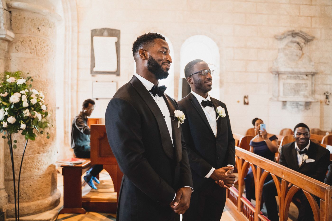 Two groomsmen stand at the altar in a beautiful church with stone walls and floral arrangements.