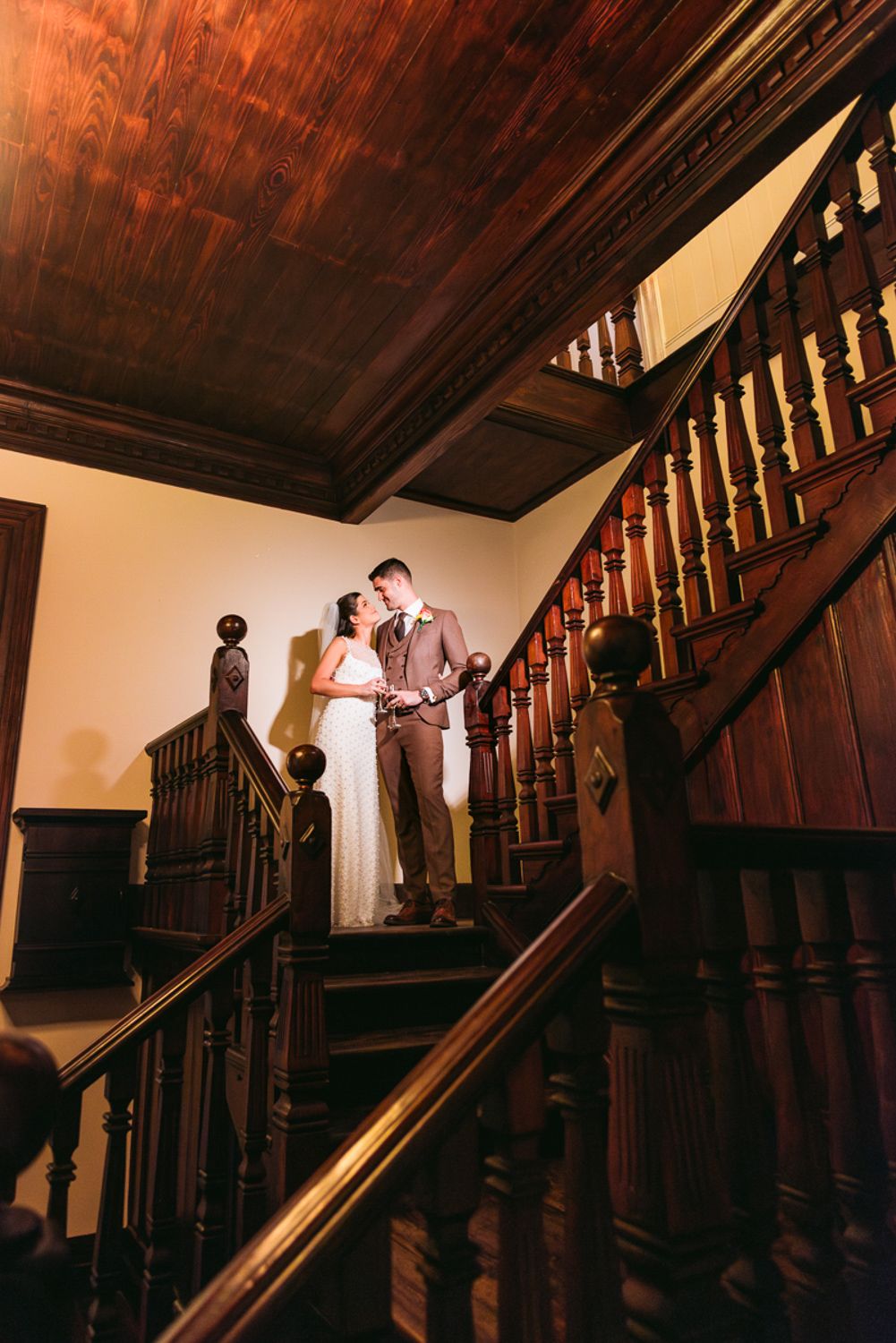 A couple in formal attire stands on an elegant wooden staircase with dark wood paneling in warm lighting.