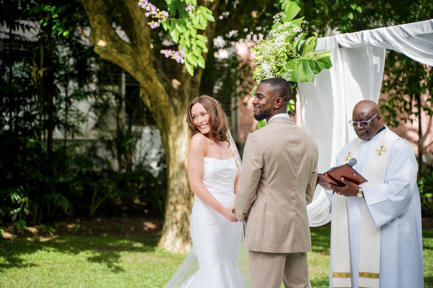 Intimate garden wedding ceremony under natural tree canopy with white fabric backdrop.