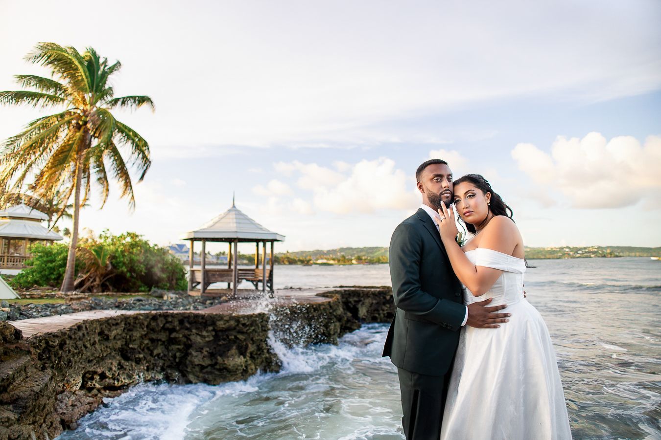 A romantic wedding couple embraces on a tropical beach with palm trees and ocean waves at sunset.