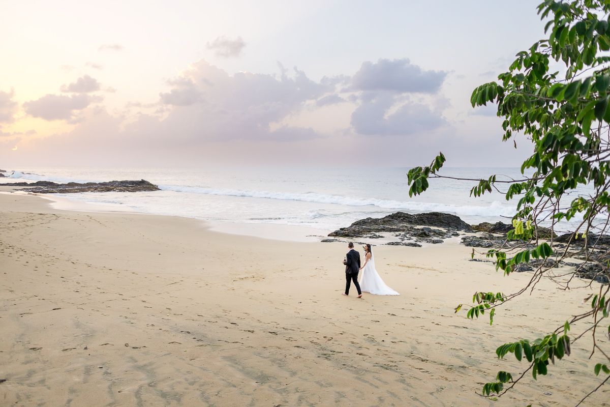Bride and groom walking on a serene sandy beach at sunset with gentle waves and pastel skies.