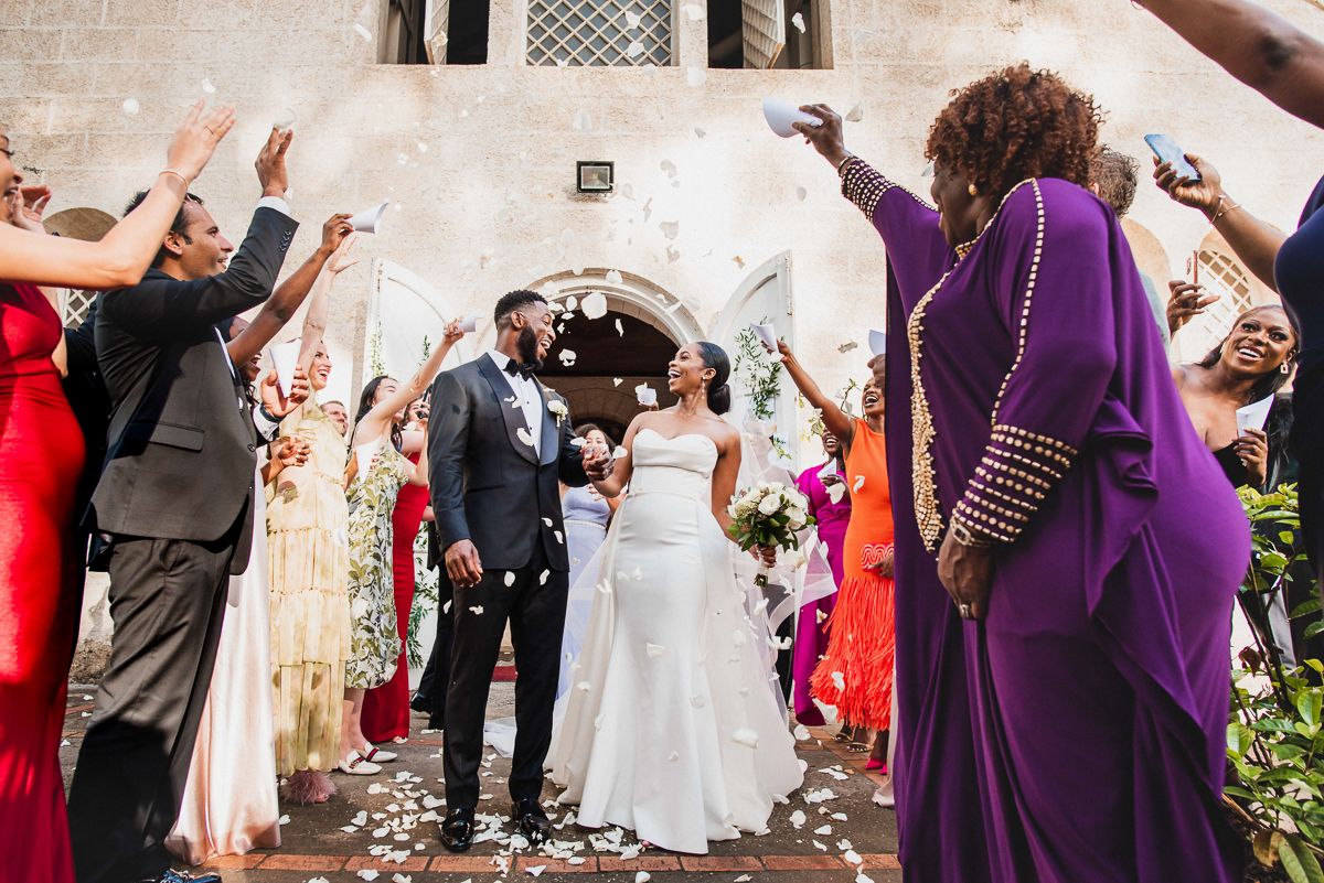 Bride and groom exit church ceremony as guests celebrate with confetti and flower petals at outdoor wedding.