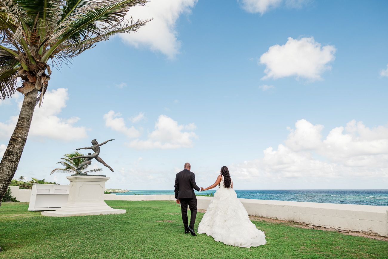 Bride and groom holding hands on oceanfront lawn beside a dancer statue at a tropical beach wedding venue.