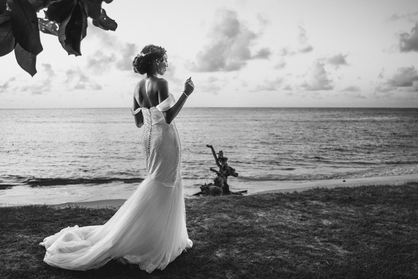 Bride in elegant white gown stands on tropical beach at sunset in black and white wedding photo.
