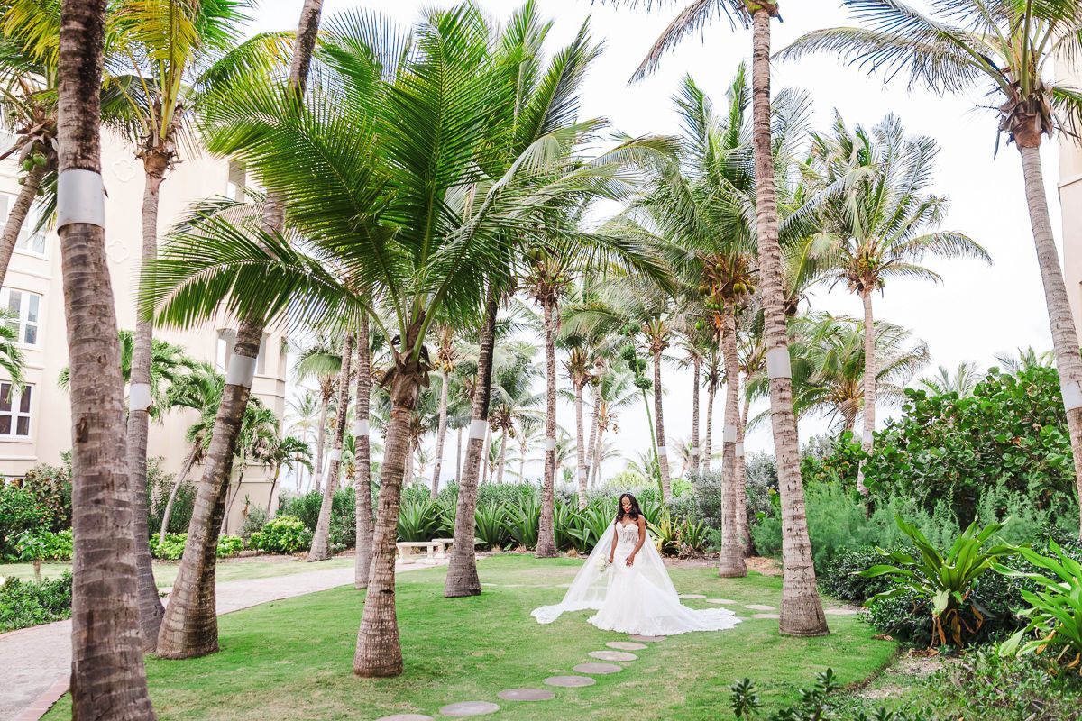 Bride in white gown poses among tropical palm trees on lush green lawn at a beachside resort wedding venue.