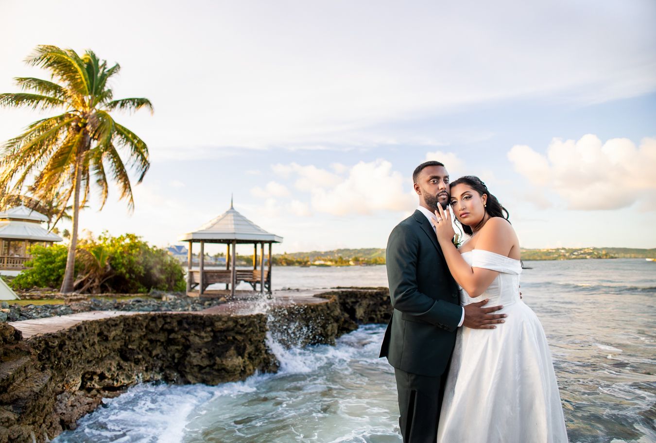 Wedding couple poses along scenic rocky coastline with palm trees and crashing waves at sunset.