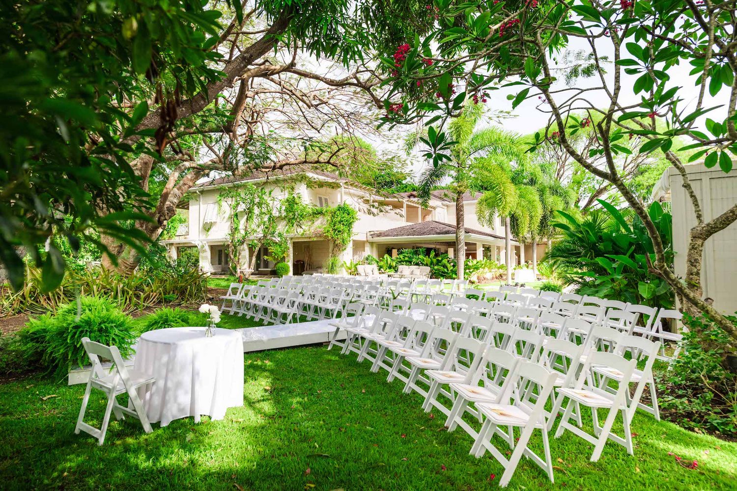 White chairs set up for outdoor wedding ceremony on green lawn with Spanish style house in background. Villa Aama in barbados