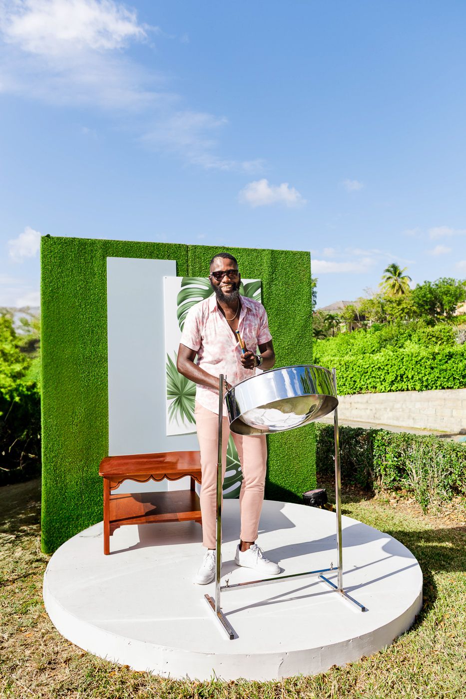 Steel drum musician performs against a tropical green backdrop on an outdoor circular stage.