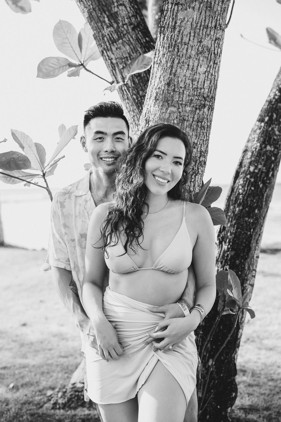 A couple embraces in black and white photography next to a tree trunk.