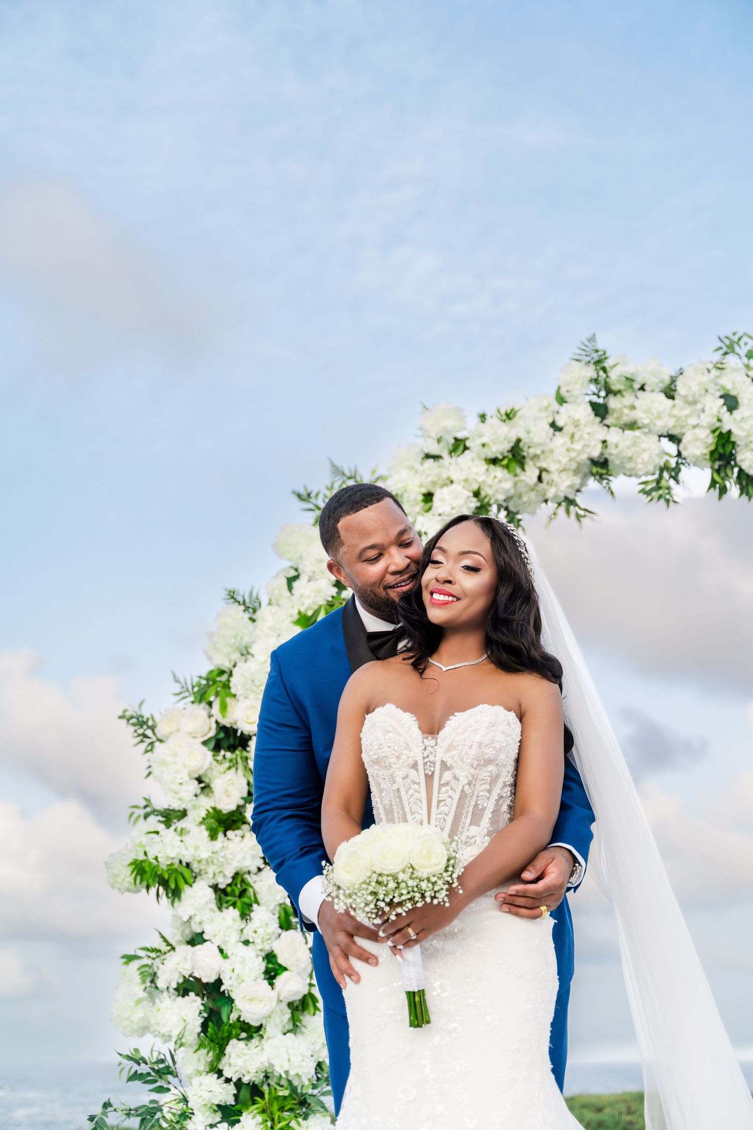Newlywed couple embraces during outdoor beach wedding ceremony with white floral arch against blue sky.
