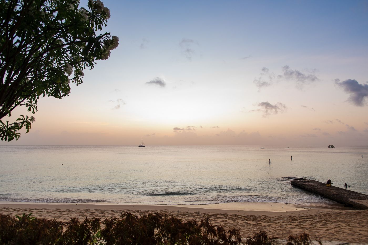 Tranquil beach sunset with wooden pier and boats on horizon.