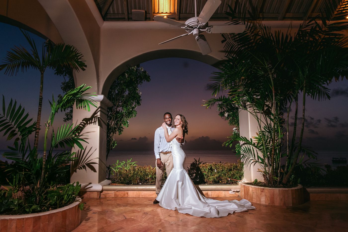 A couple shares a romantic dance moment on an elegant terrace with palm trees and arched architecture at sunset.