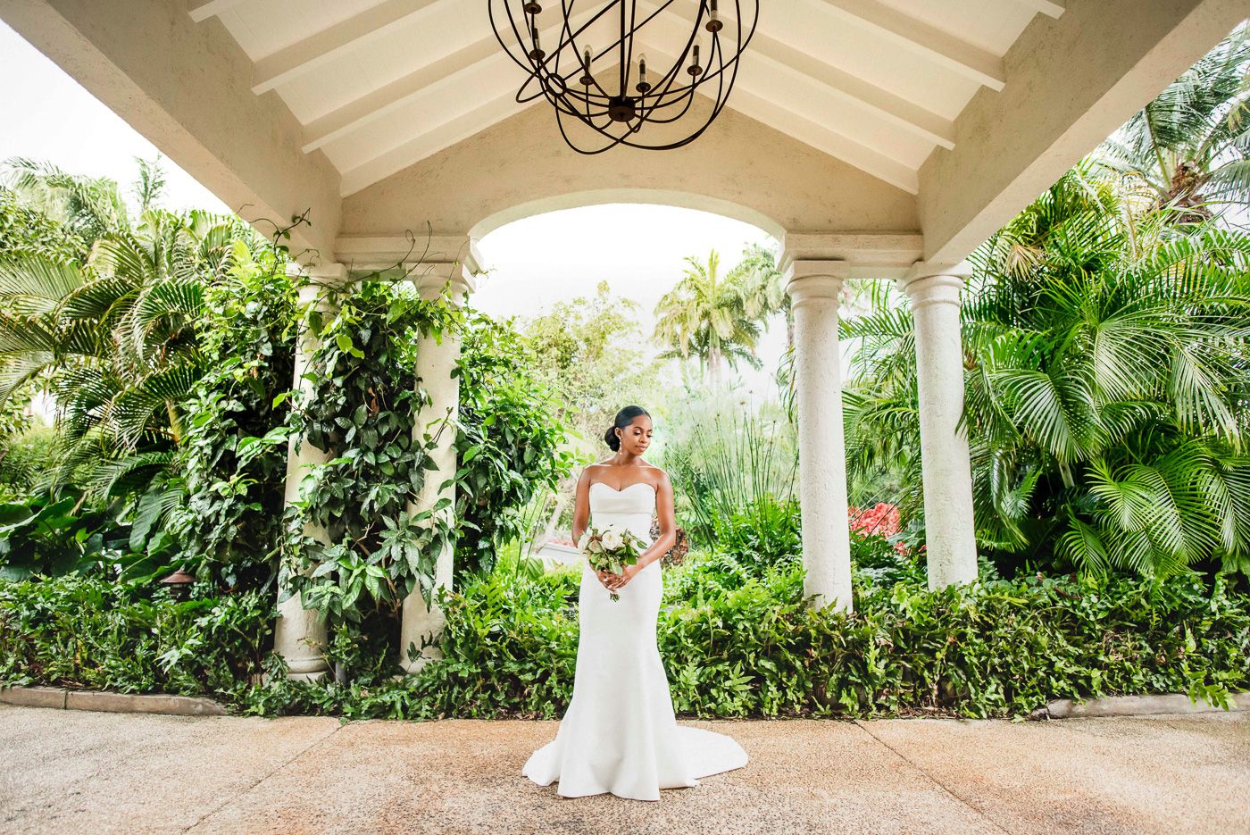 Bride in elegant white gown stands alone under covered portico surrounded by lush tropical gardens.