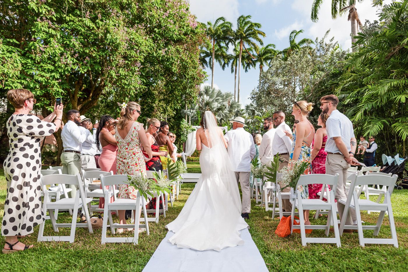 Wedding ceremony in progress with guests seated on white chairs along a white aisle runner in a garden.
