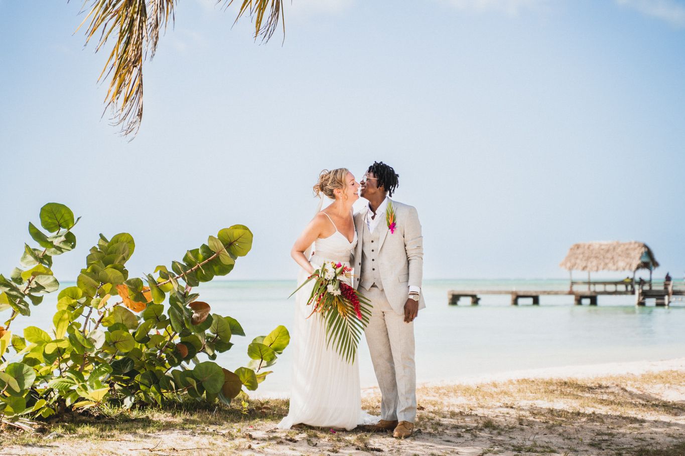 A romantic beach wedding ceremony with a couple embracing on a tropical shore with an overwater bungalow in the background.