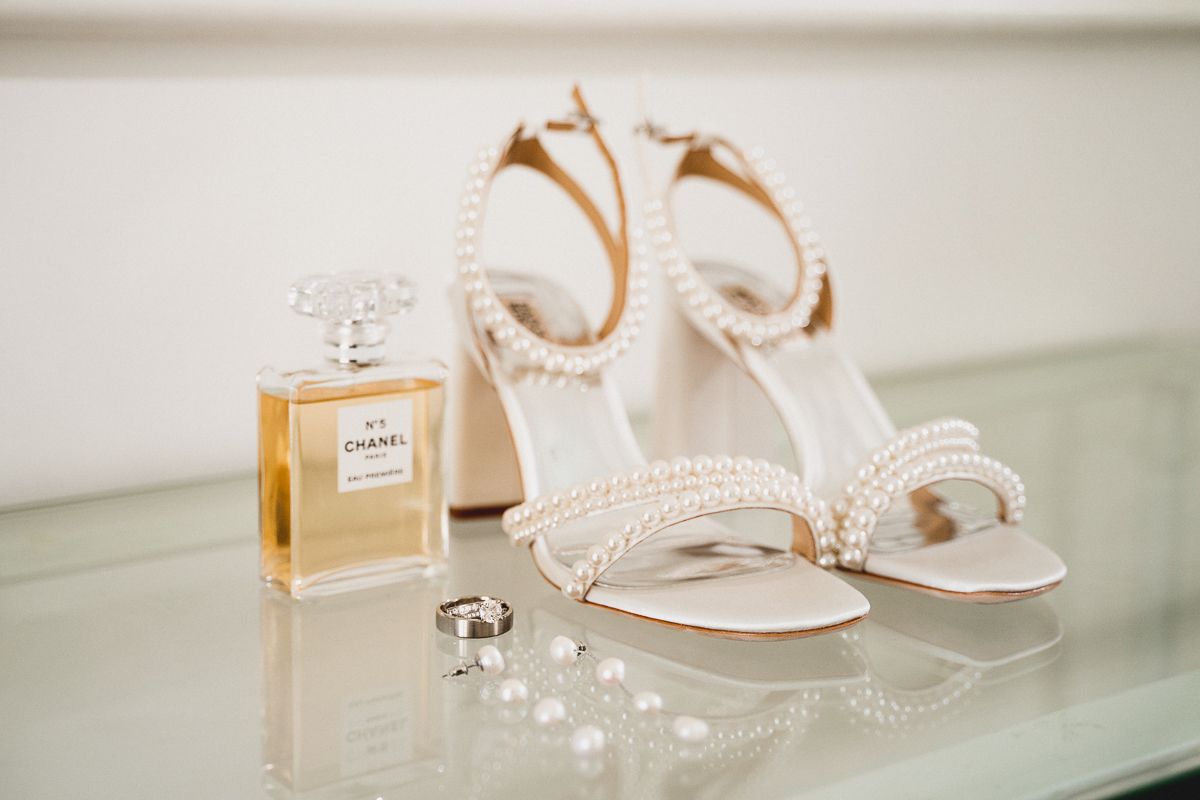 Pearl-adorned bridal heels beside Chanel No.5 perfume and wedding rings on a glass surface.