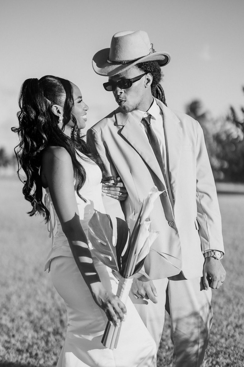 Black and white photo of an elegant couple in formal attire, man in suit and cowboy hat, woman in white dress outdoors.
