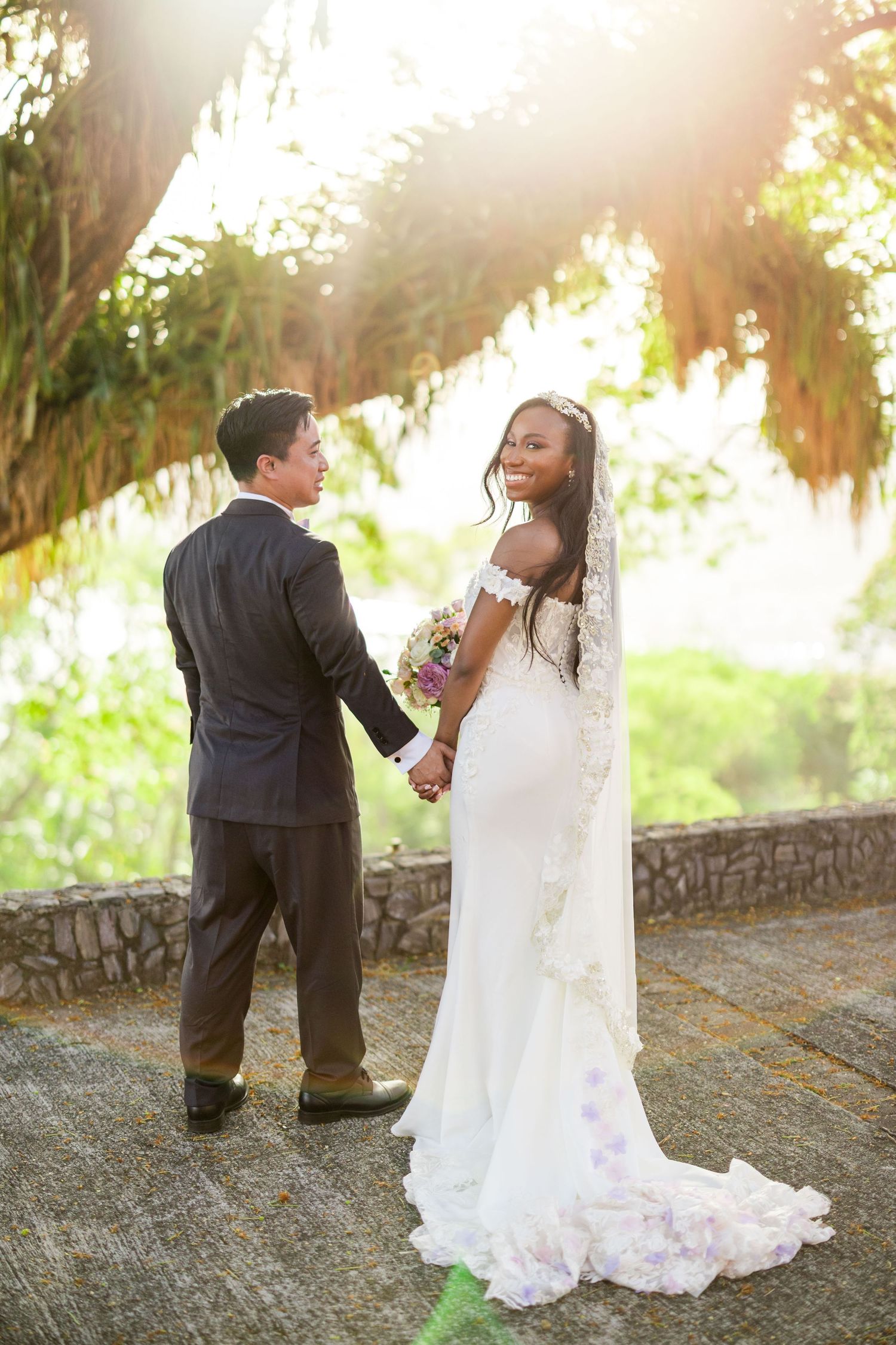 Bride in white gown holding bouquet smiles back at groom in black suit, holding hands outdoors in golden sunlight.