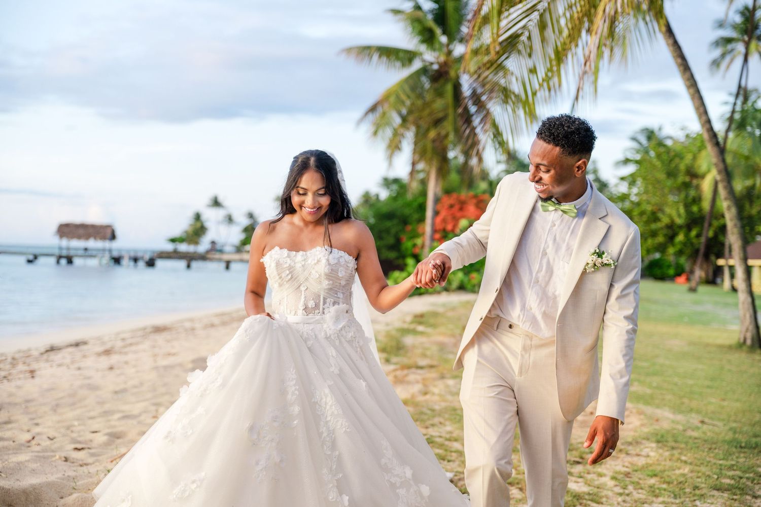 Bride in white lace gown and groom in beige suit hold hands walking on tropical beach with palm trees.