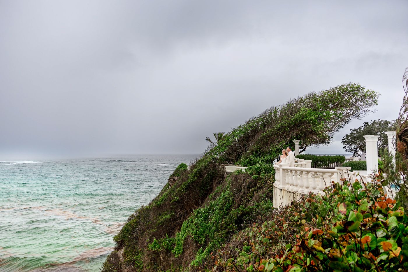 Stormy coastal cliffside with lush greenery, white balustrade, and turbulent ocean waves under overcast skies.
