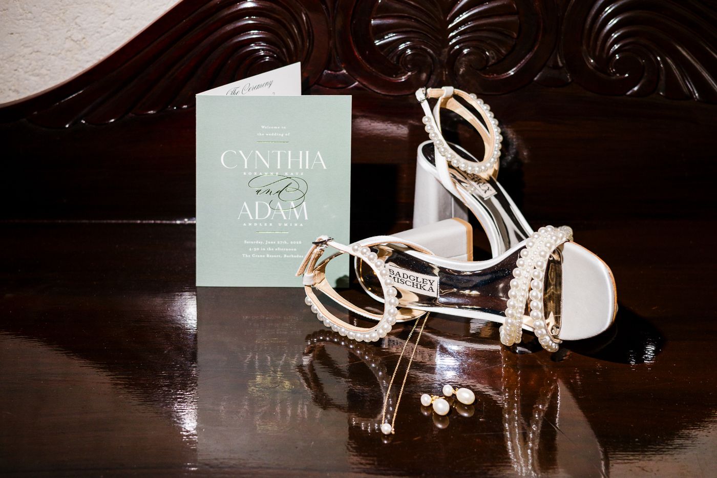 Elegant white strappy heels with pearl details beside a mint green Cynthia and Adam wedding invitation on a wooden surface.