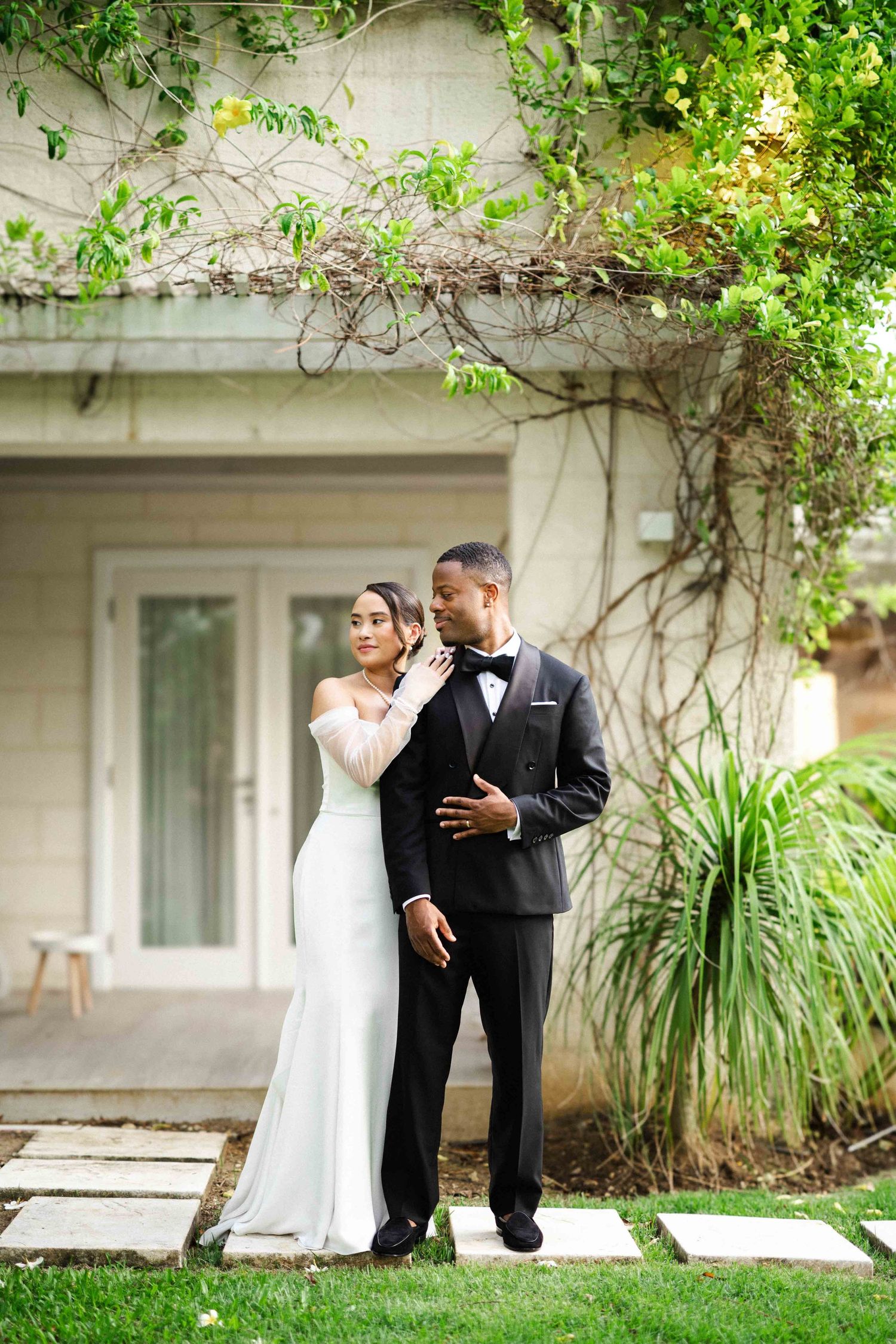 A romantic wedding couple embraces in a garden setting with lush greenery and white walls.