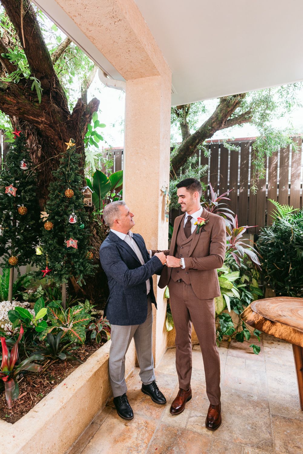 Two people share a heartfelt moment on a covered patio with lush greenery and plants in the background.