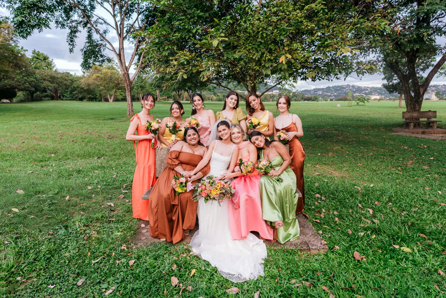Wedding party in coral dresses poses together on a grassy hillside during an outdoor celebration.