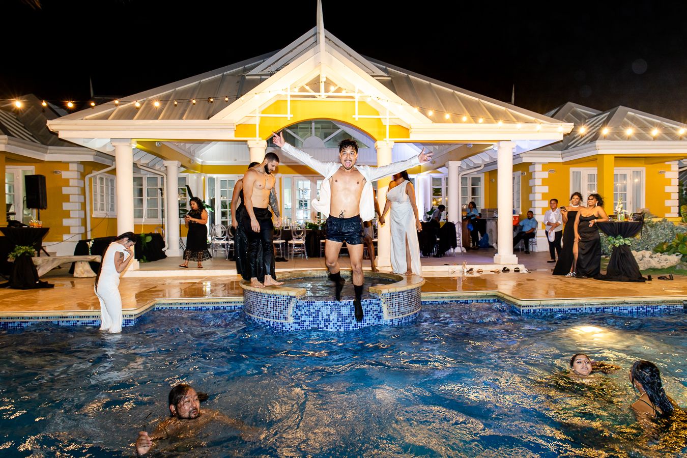 Wedding guests jumping into illuminated pool with white gazebo backdrop at night.