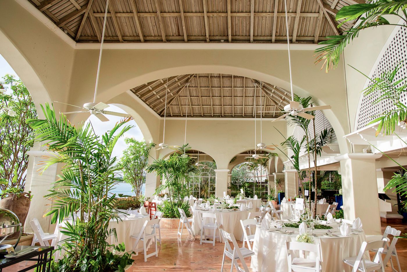 Elegant wedding reception venue with white decor and tropical palm trees under vaulted ceiling.