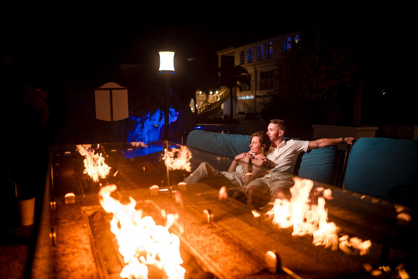 Couple relaxing on outdoor furniture next to dramatic fire features at night.