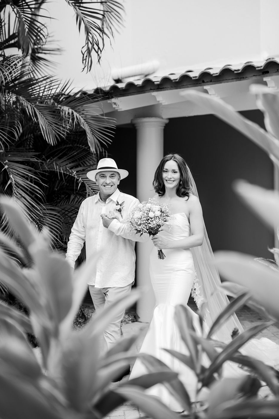 A tropical wedding scene with palm trees framing a couple walking along a Spanish-style building.