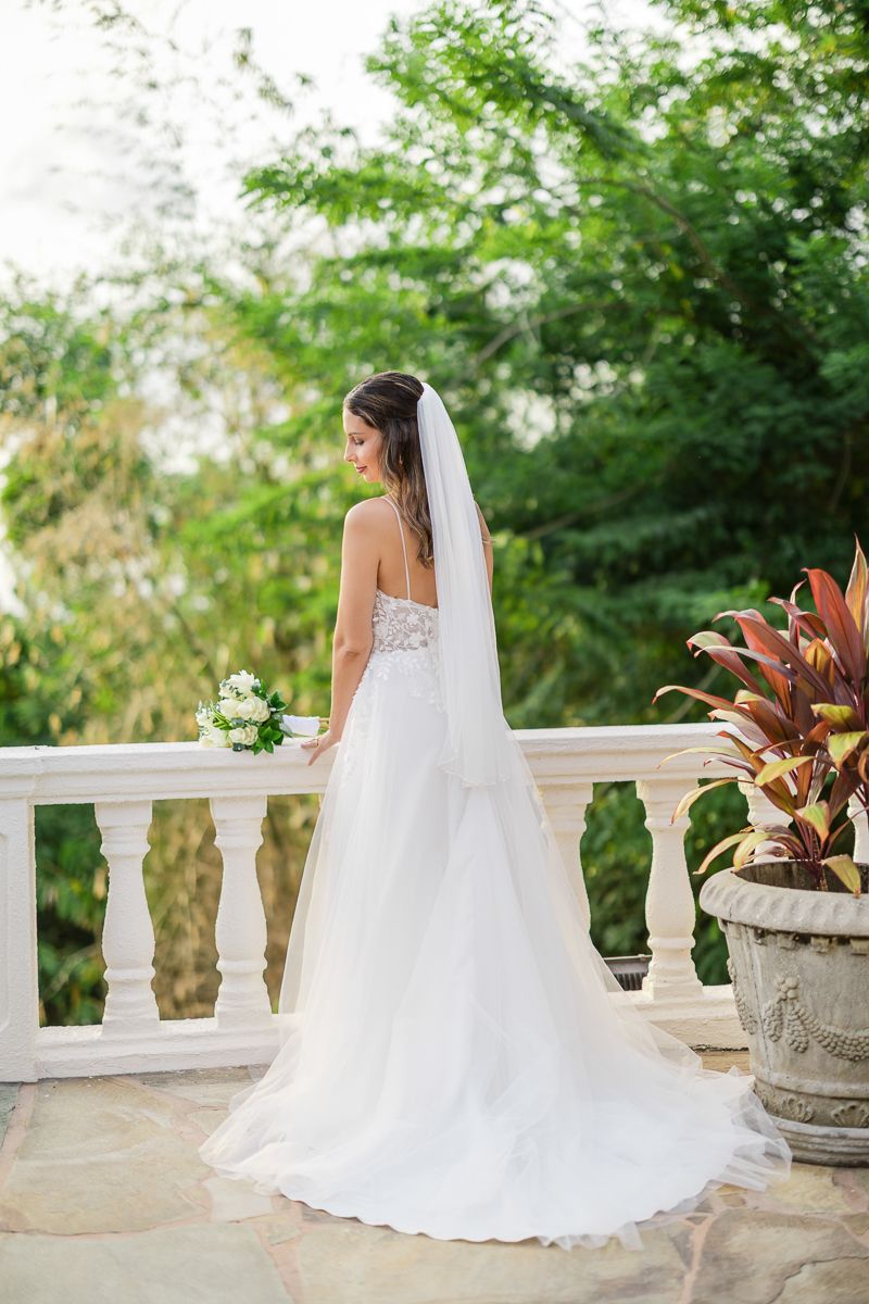 Bride in white strapless gown with veil holding bouquet on elegant balcony surrounded by lush greenery.