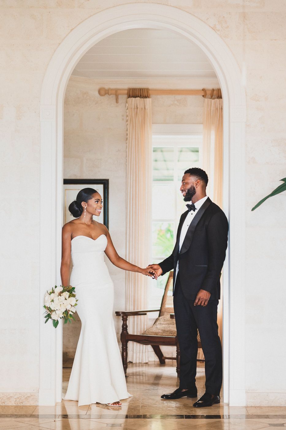Elegant bride in strapless white gown holds bouquet while groom in black tuxedo holds her hand under a white arch.