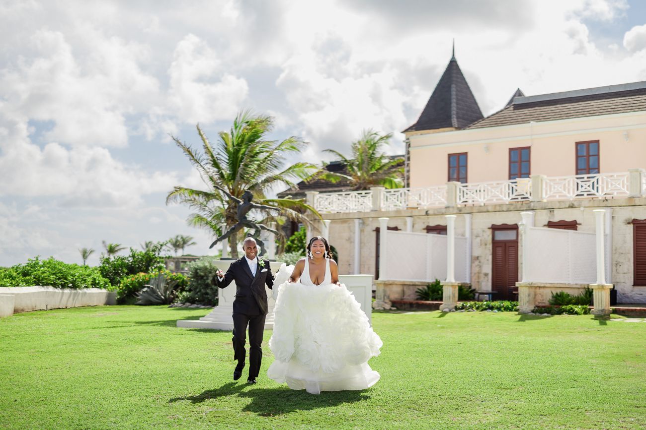 Joyful newlyweds celebrate on lush green lawn outside elegant estate venue with palm trees and cloudy sky.
