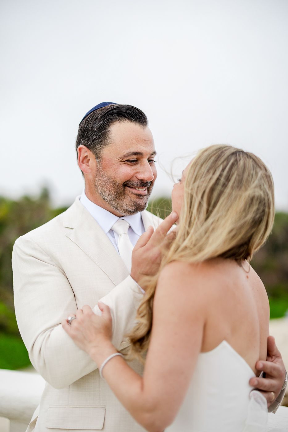 Groom in cream suit tenderly touches bride's face during romantic outdoor wedding ceremony.