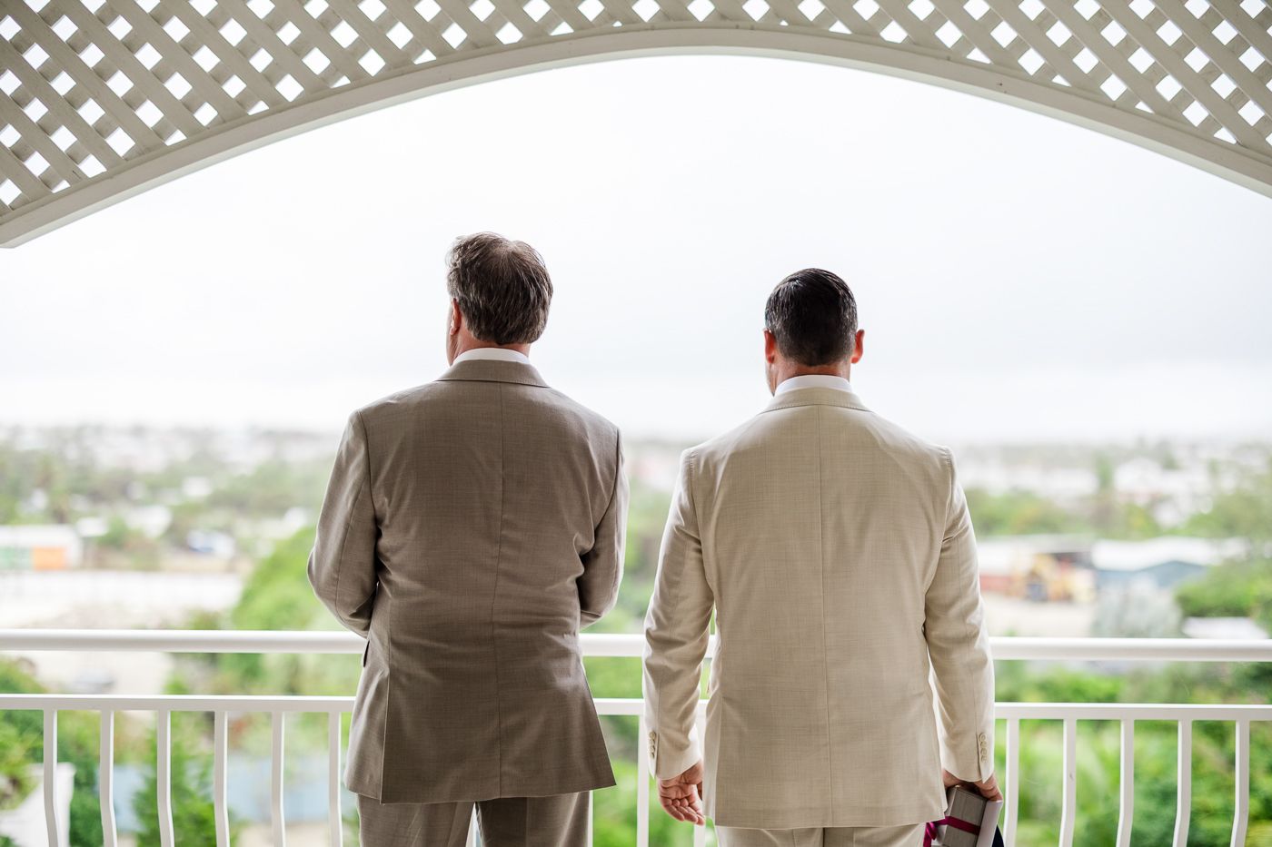 Two men in beige suits stand on a balcony overlooking a city skyline.