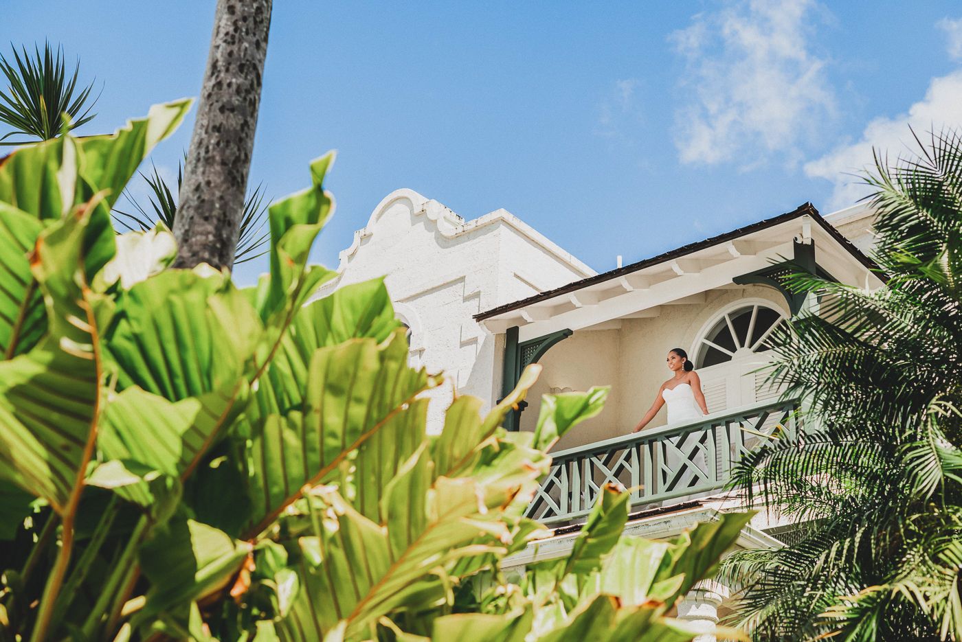 Tropical palm trees and banana plants frame a white Caribbean-style building with a balcony under bright blue skies.