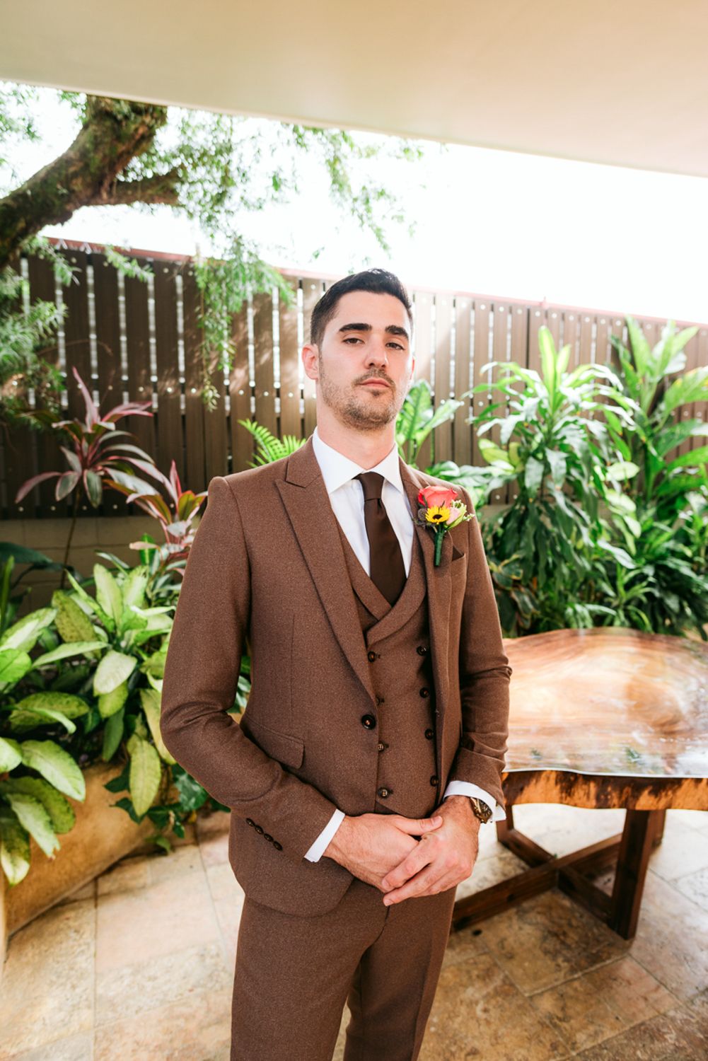 A formal brown three-piece suit is displayed in outdoor garden setting with lush tropical plants and wooden fence backdrop.