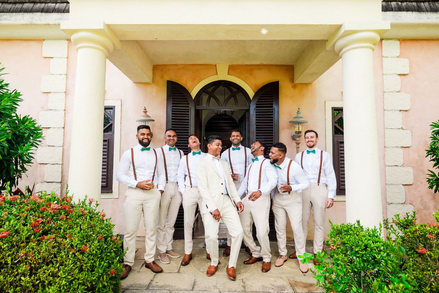 Groomsmen in white shirts and suspenders pose together outside a Mediterranean-style building.