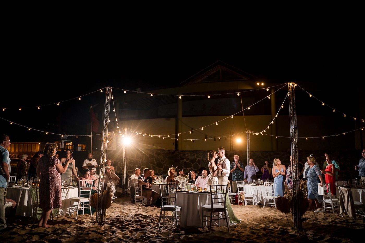 Panoramic view of a nighttime beach wedding reception with twinkling lights.