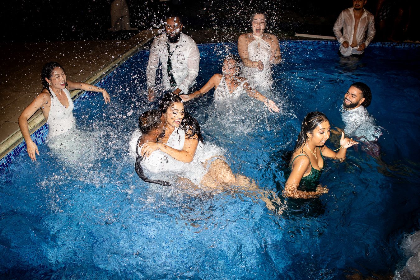 Wedding party creates splashes in pool during nighttime celebration.