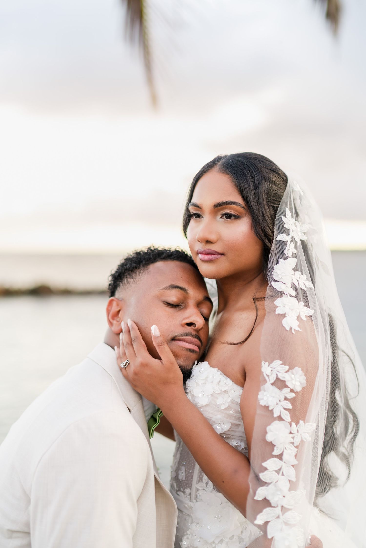 A bride in a lace veil and gown embraces her groom in a beige suit during a romantic beach wedding ceremony.