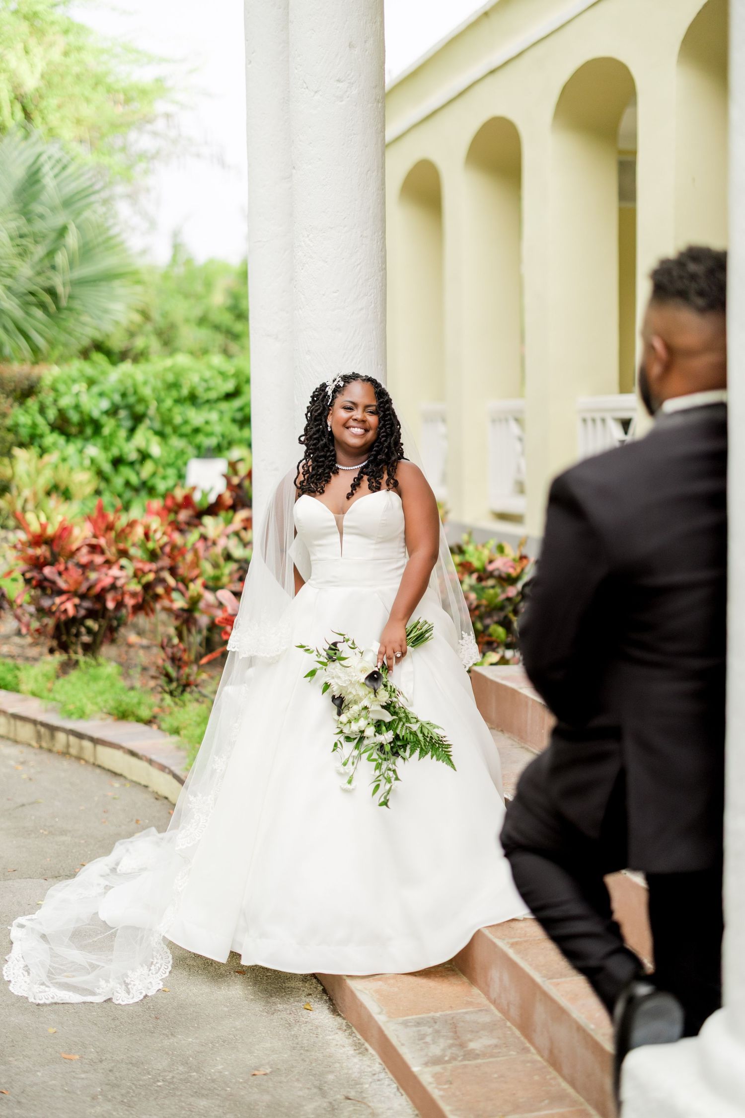 Bride in white strapless ball gown with veil holds bouquet by column, smiling at groom in black suit outdoors.
