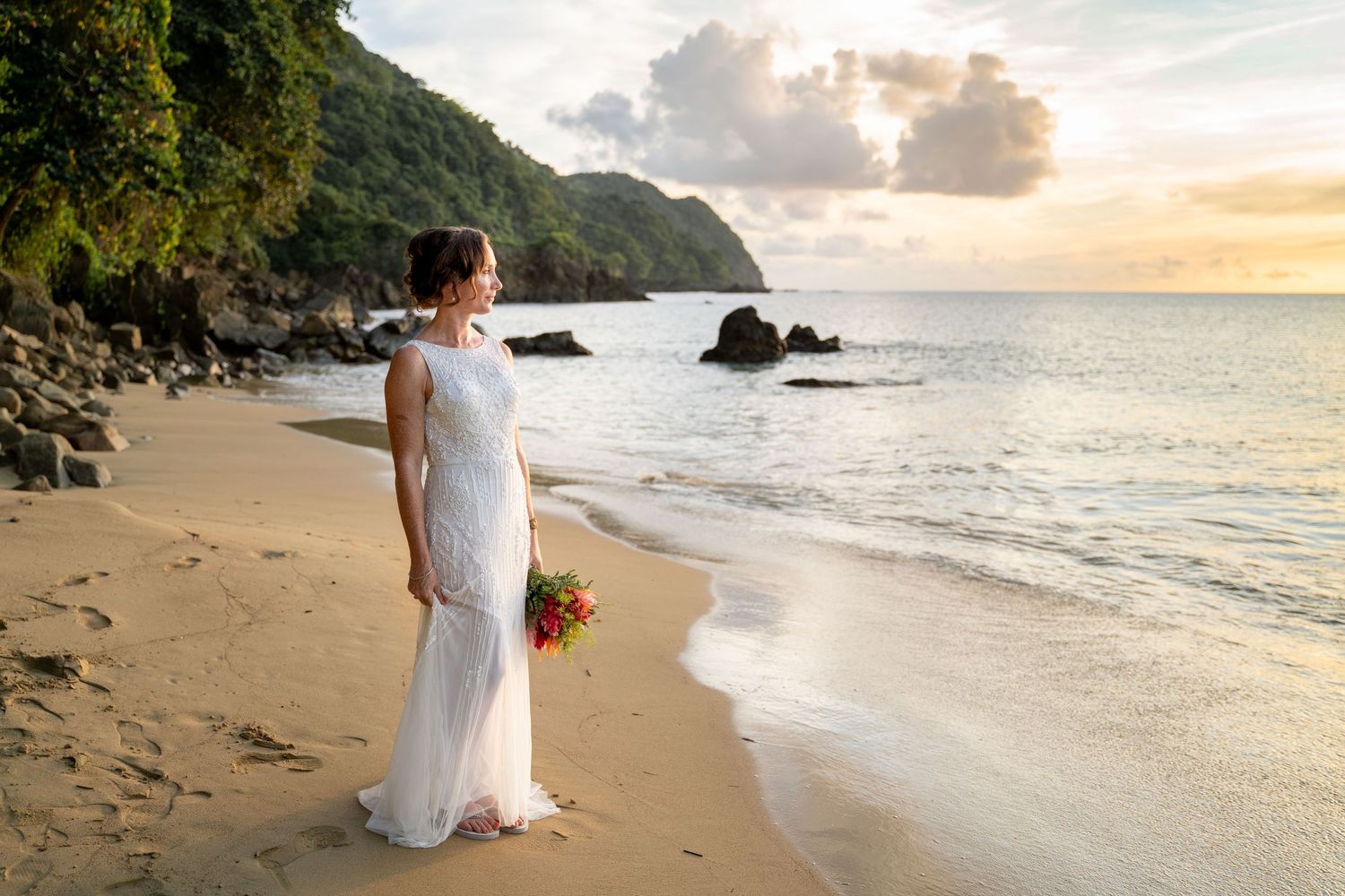 Bride in white gown holding bouquet on tropical beach at sunset with rocky coastline.