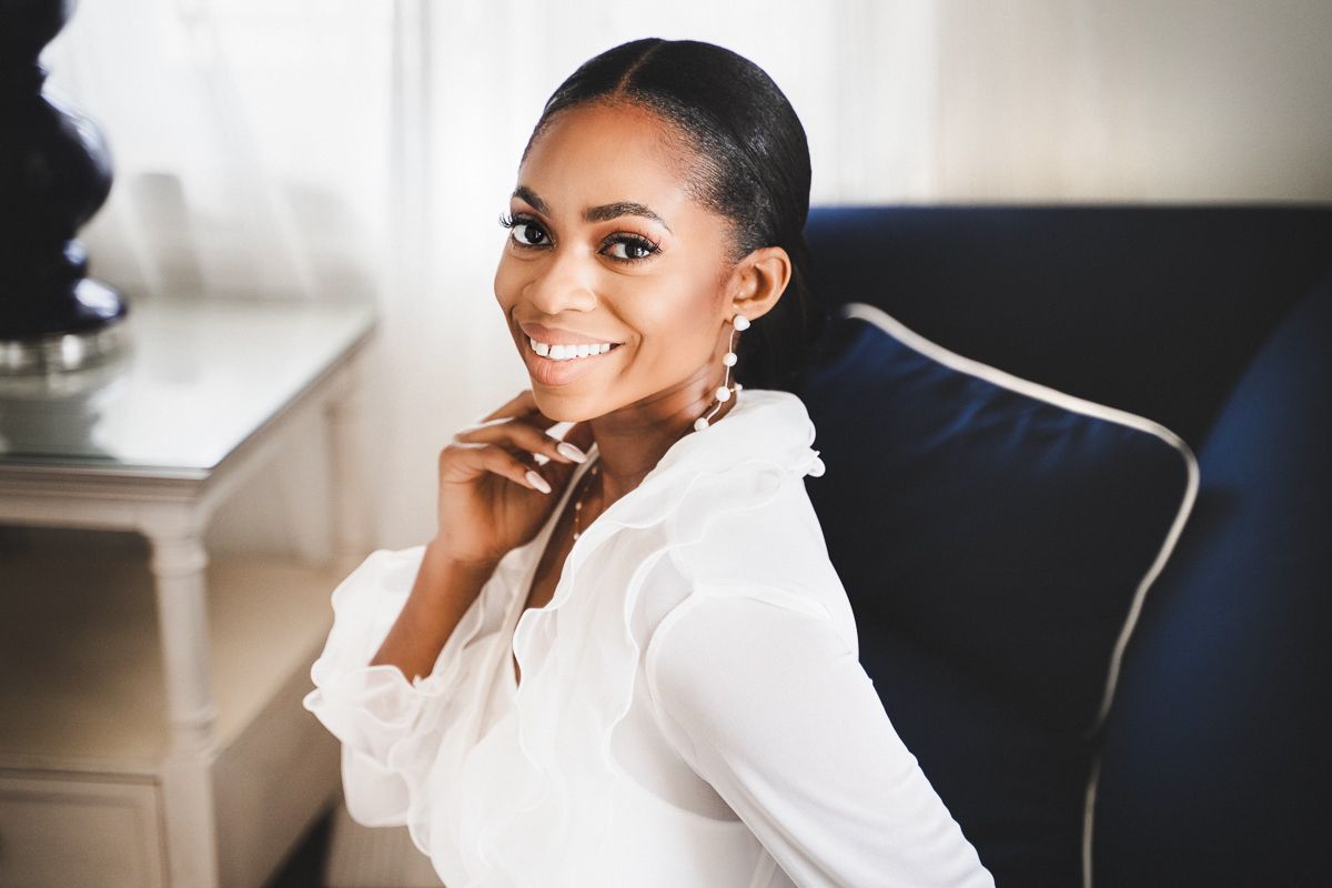 Smiling young Black woman in white blouse seated in blue chair, wearing earrings, posing elegantly indoors.