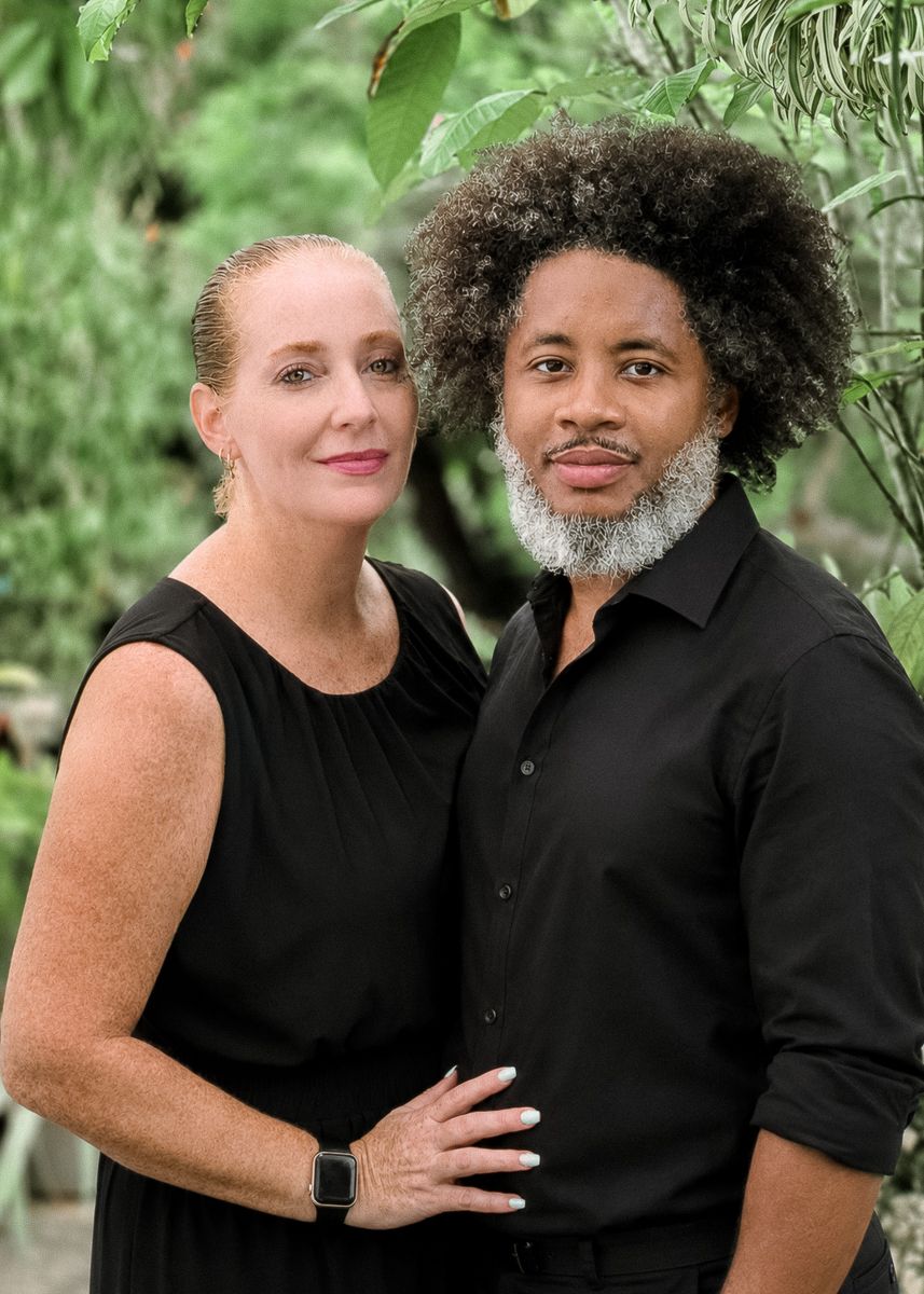 A couple in matching black outfits posing together outdoors surrounded by lush green foliage.