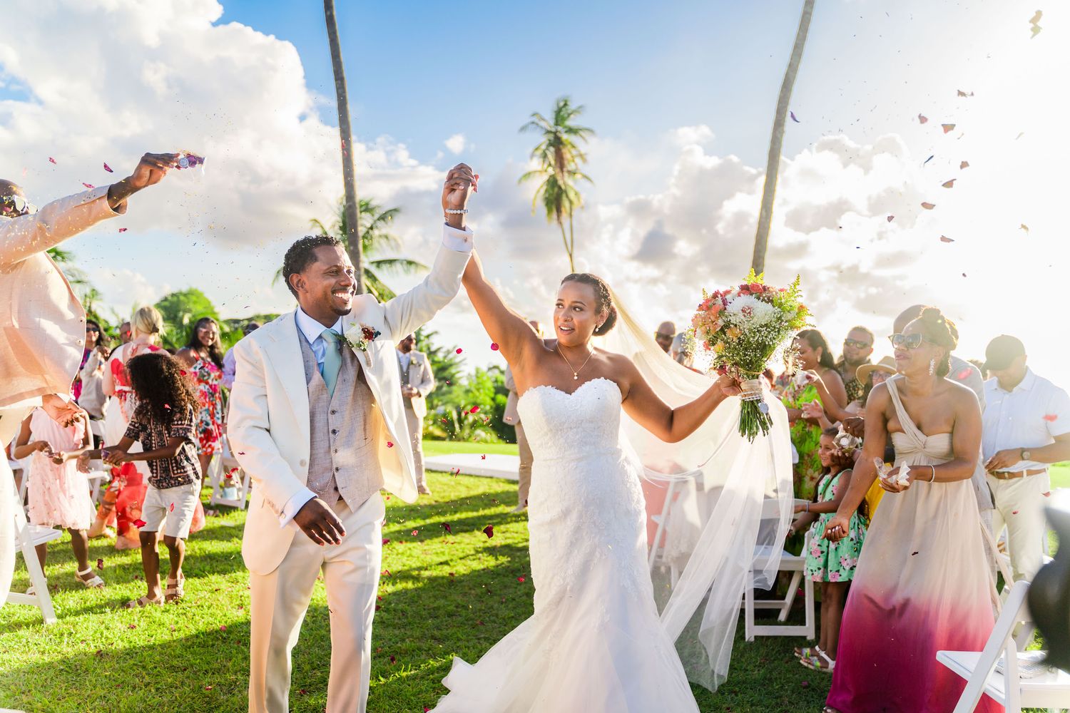 Joyful newlyweds celebrate outdoor tropical wedding ceremony with guests tossing colorful flower petals under blue sky.