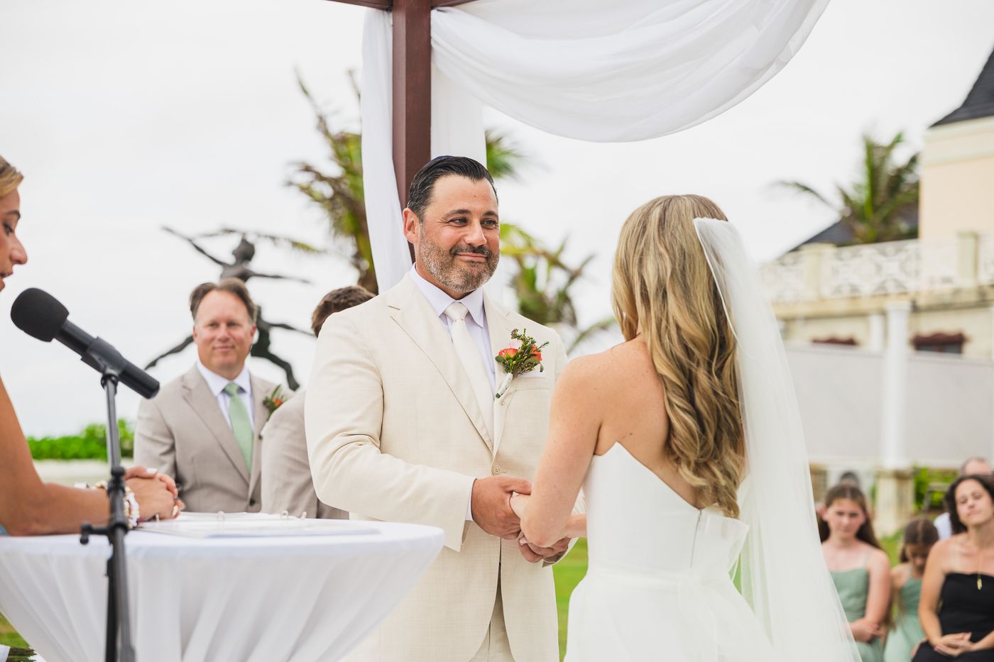 Groom in white suit smiling at bride during outdoor wedding ceremony with tropical backdrop.