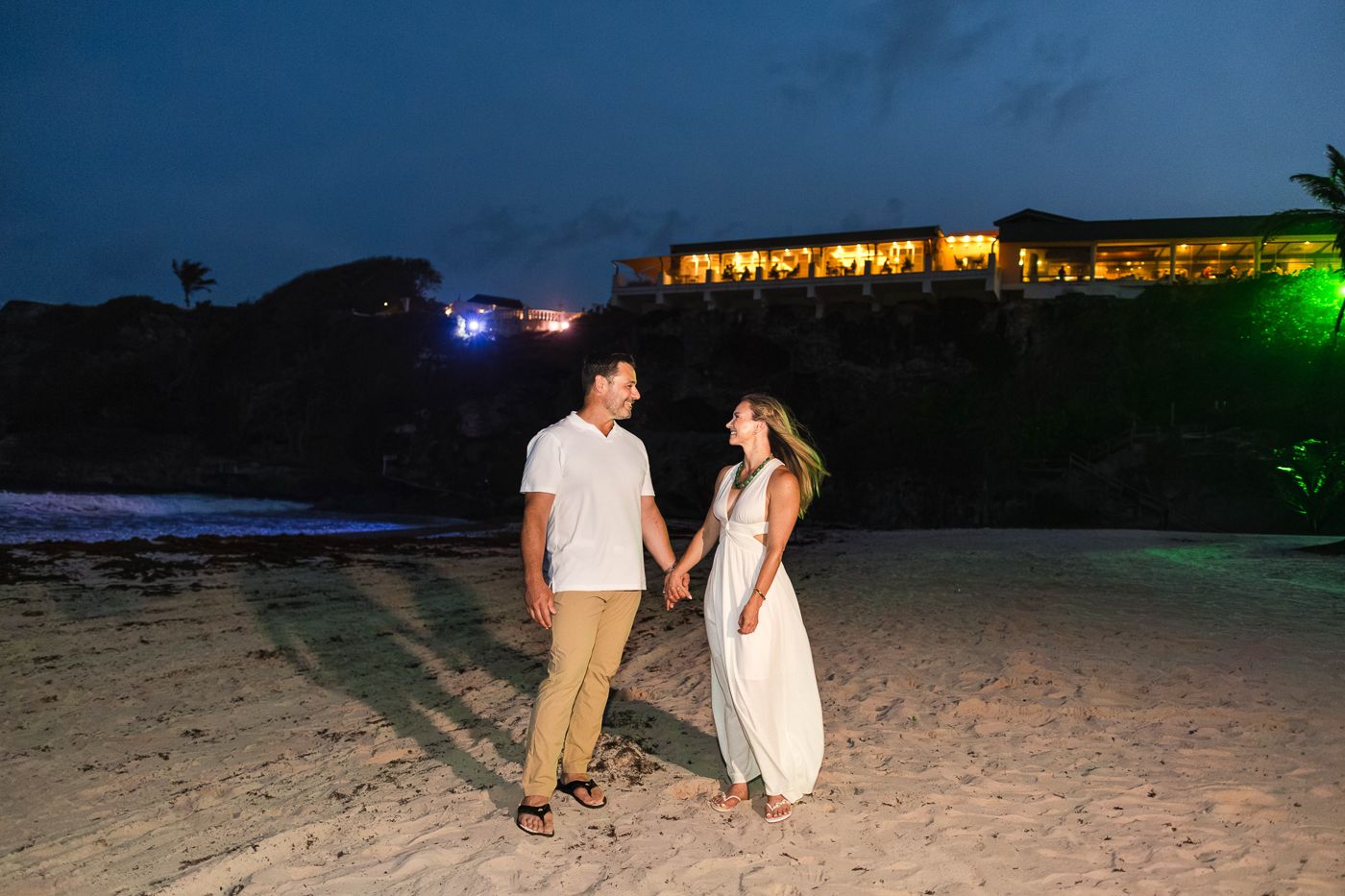 Couple holding hands on a sandy beach at dusk with illuminated cliffside resort in background.
